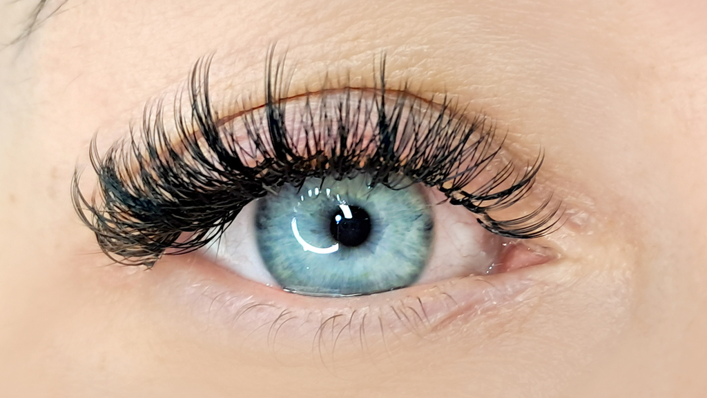 A woman with bright blue eyes and a Doll style lash extension. The doll style eyelash extension is thick and becomes thicker and longer towards the outside of her eye.