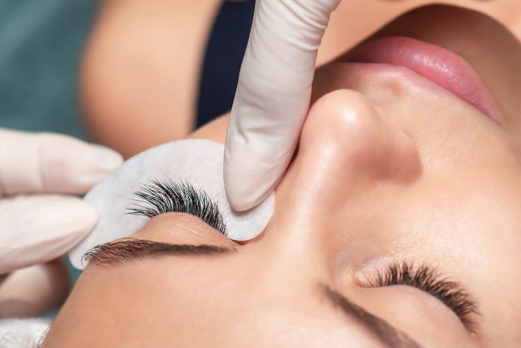 A client lying down with eyes closed during a lash appointment. A lash artist wearing gloves applies classic eyelash extensions. One eye has lash extensions with a pad underneath. The other eye has natural lashes. This shows a classic eyelash extension style.
