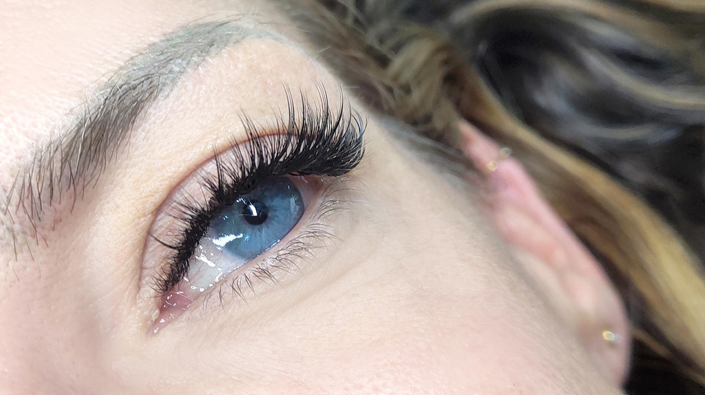 A close up of a woman on her blue eye with black hybrid lash extensions. The lashes are much fuller on the top lash and are longer towards the outside. She has very fair eyebrows.