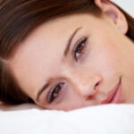 A woman with brown hair and eyes laying on a pillow with eyelash extensions.
